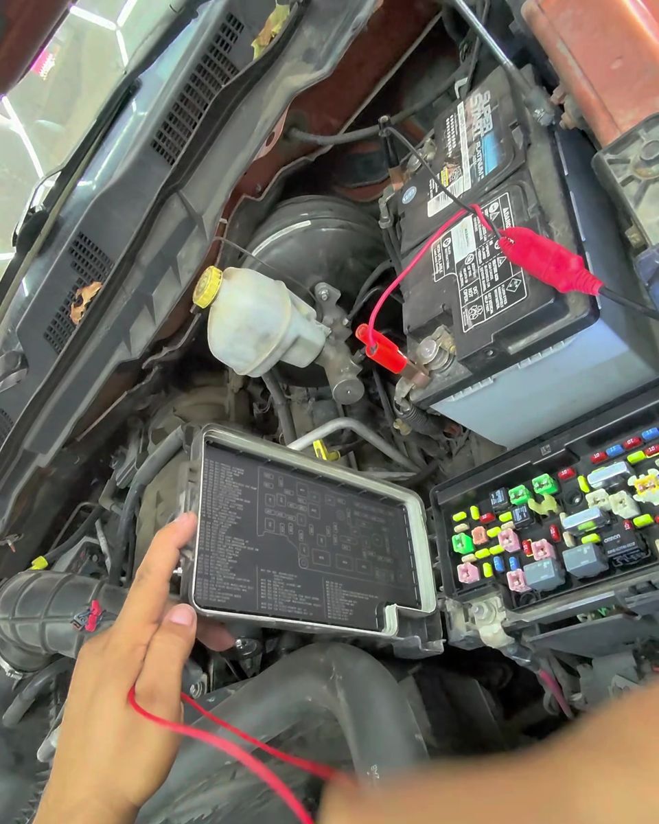 A hand holding test leads beside an open under-hood fuse box, with clips connected to the battery